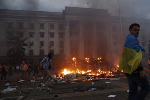 A protester wrapped in a Ukrainian flag walks past a burning tent camp and a fire in the trade union building in Odessa